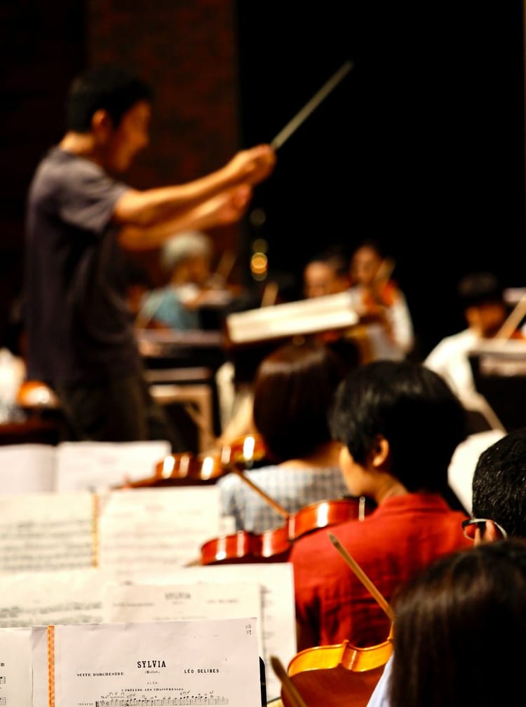 A conductor stands in the background, with three rows of string instruments in front of them.